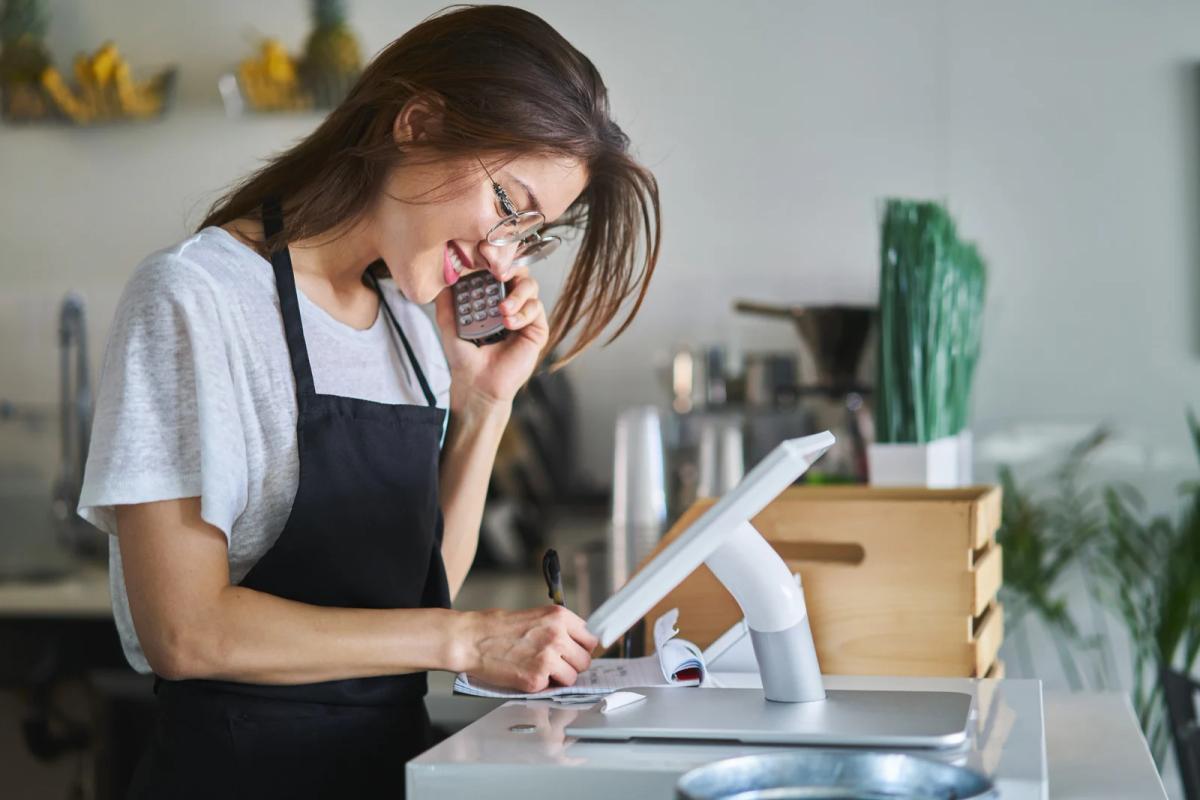 Cafe worker in apron taking notes while talking on the phone at a checkout counter