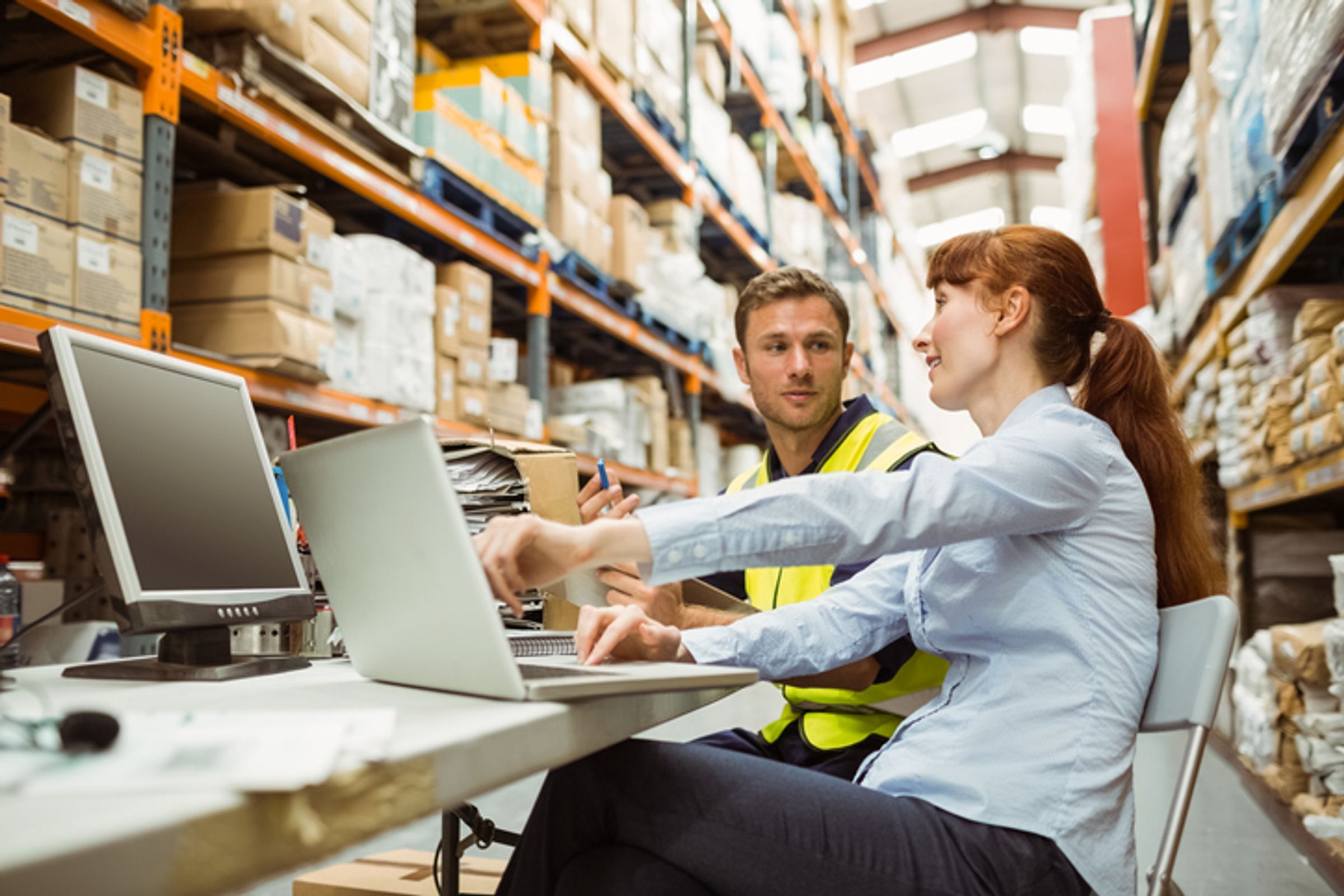 Two coworkers in a warehouse discuss data at a desk with laptops and shelves of boxes behind them