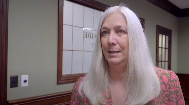 Older woman with long white hair in a pink patterned blouse standing in an office hallway
