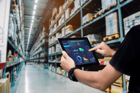 Person using a tablet with logistics dashboard in a large warehouse aisle of tall shelves