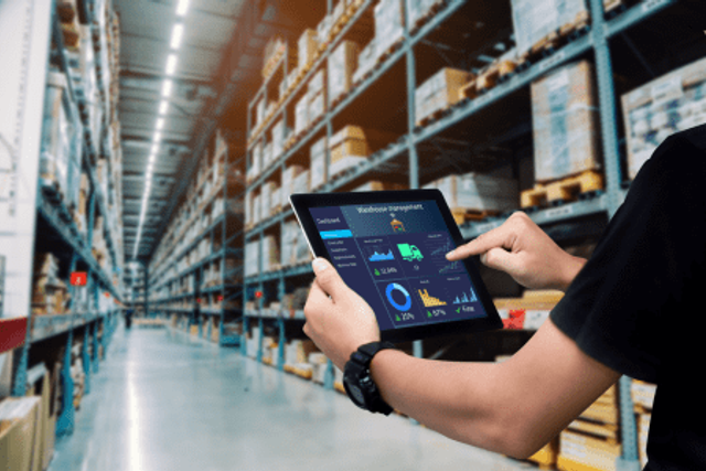Person using a tablet with logistics dashboard in a large warehouse aisle of tall shelves