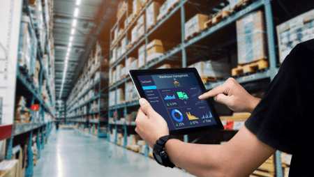 Person using a tablet with logistics dashboard in a large warehouse aisle of tall shelves