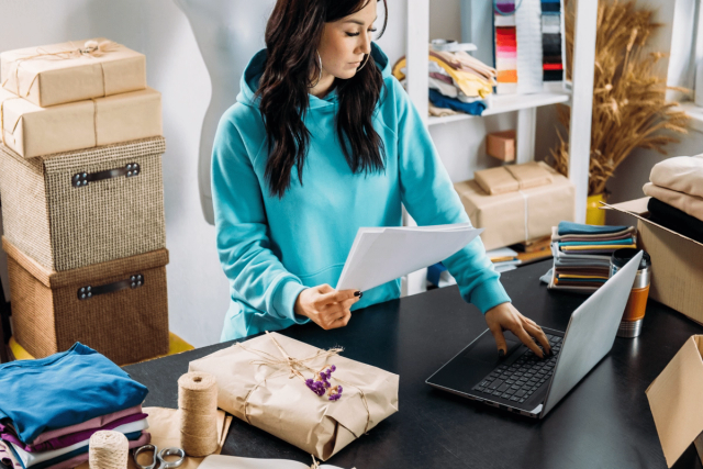 Woman in turquoise hoodie managing online orders at a packing table with parcels and fabric stacks