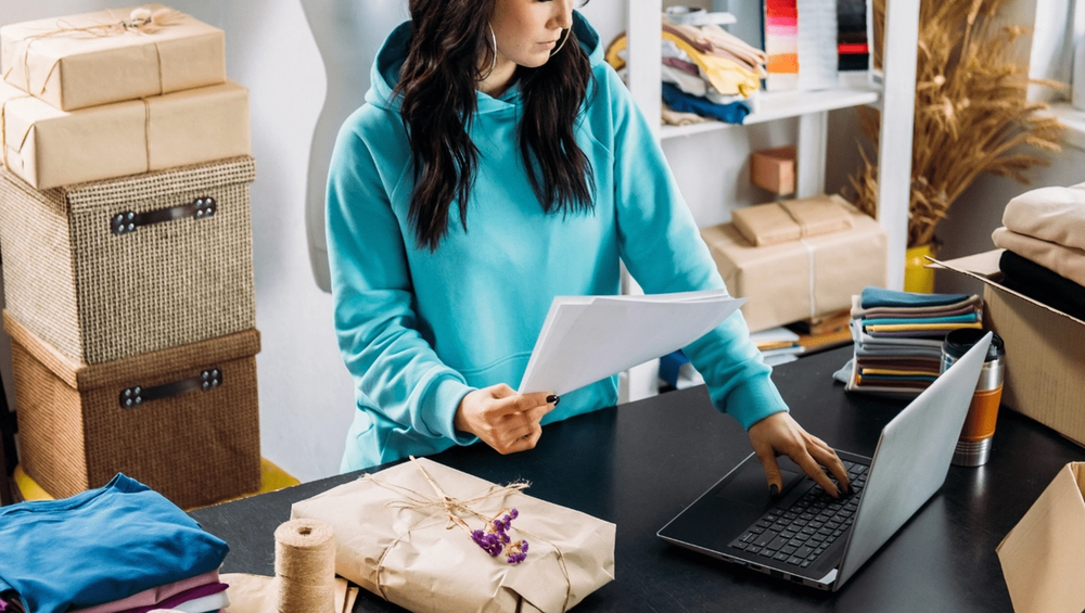 Woman in turquoise hoodie managing online orders at a packing table with parcels and fabric stacks