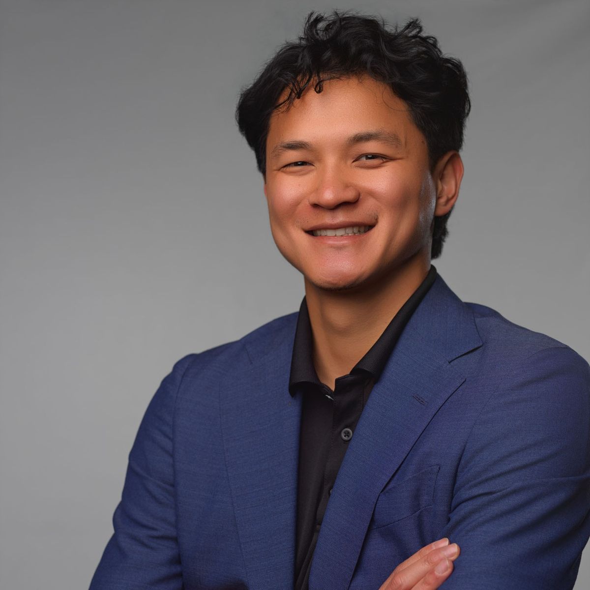 Smiling man in a blue blazer and black shirt posing against a plain gray background