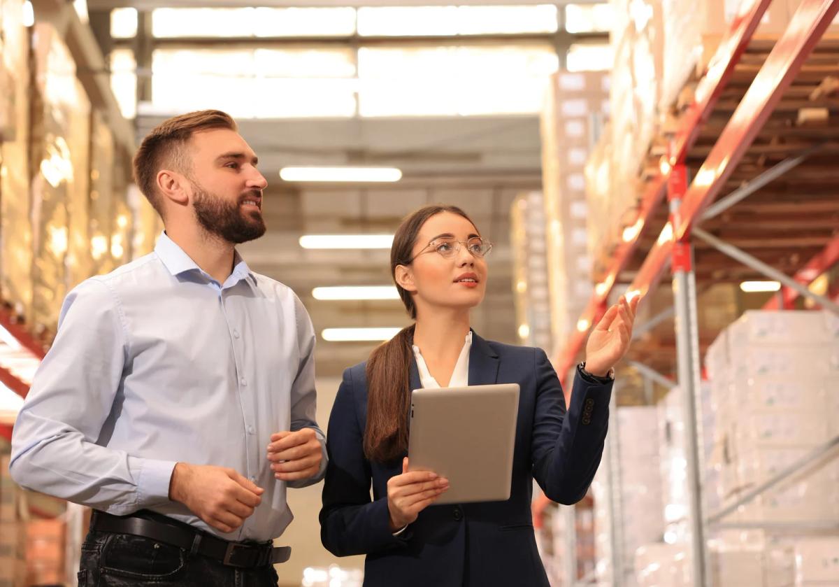 Two colleagues inspect shelves in a warehouse, one holding a tablet and gesturing toward stacked boxes.