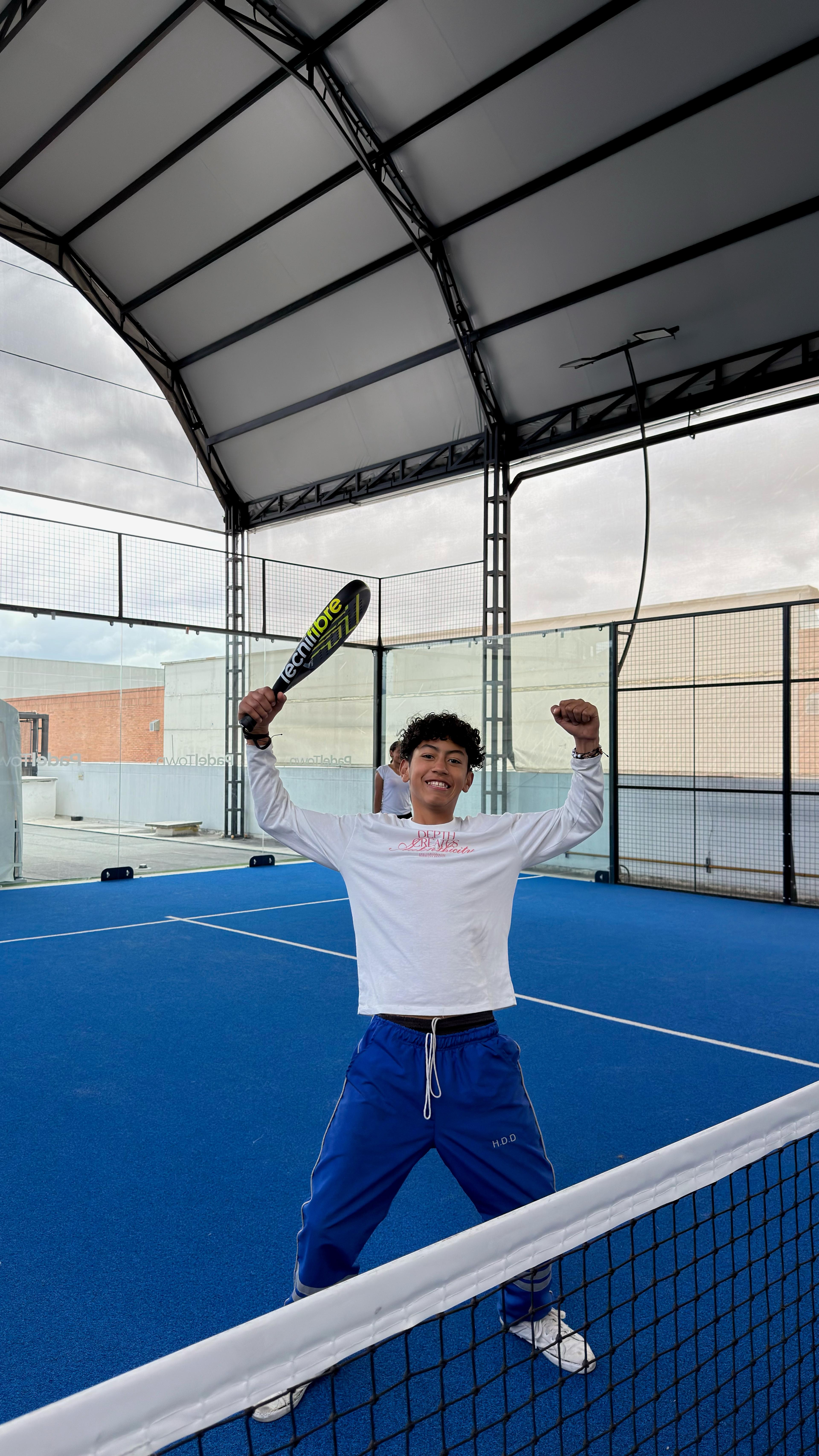Un estudiante celebra con los brazos en alto y la raqueta en la mano, junto a la red de una cancha cubierta.