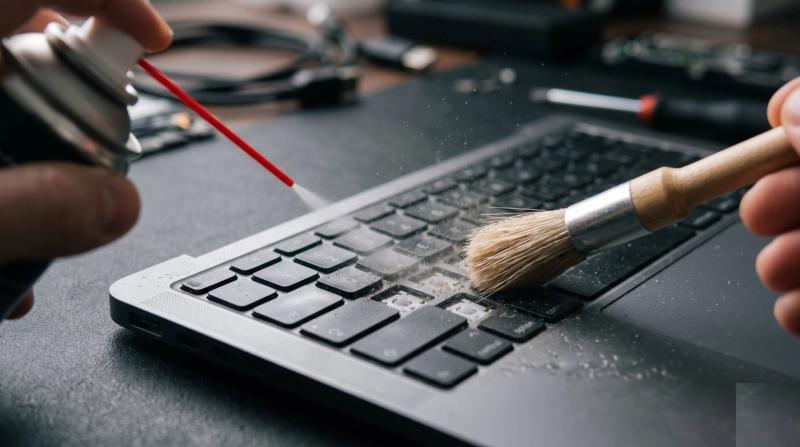 Hands using compressed air and a soft brush to clean dust out of a laptop keyboard on a repair bench