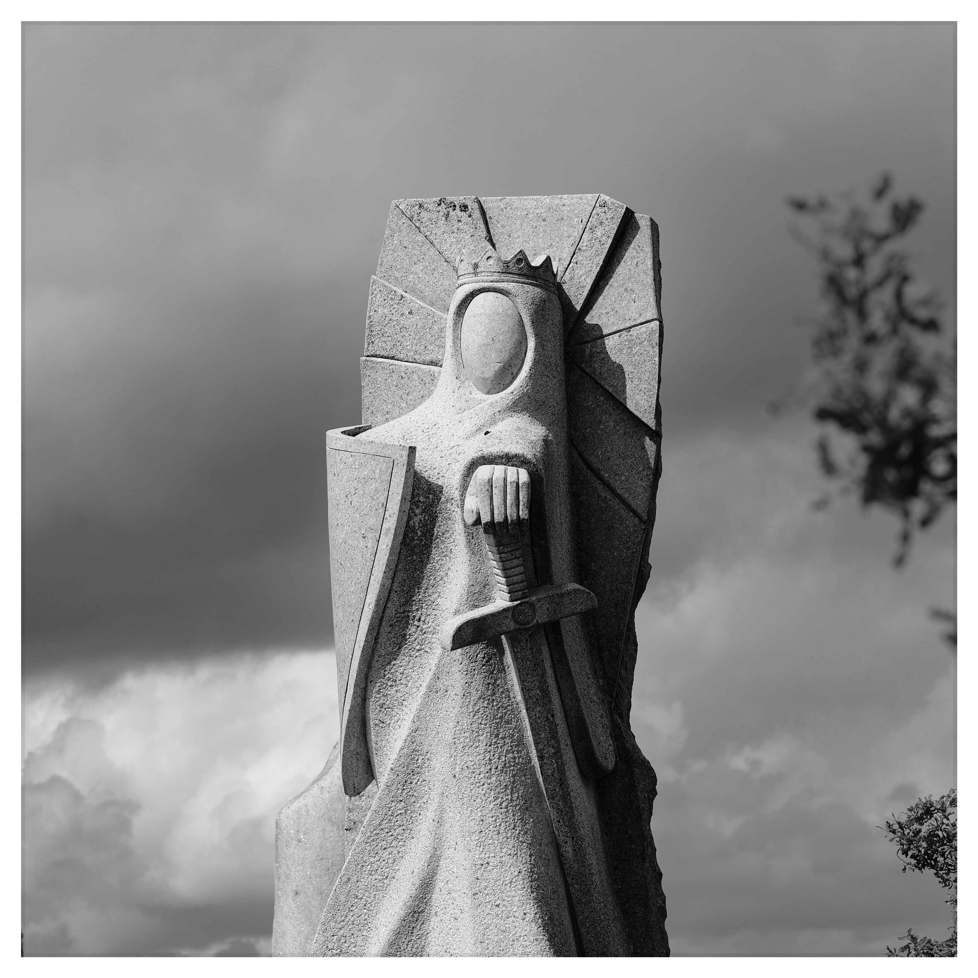 Black-and-white photograph of a monumental stone sculpture of a crowned figure holding a sword, seen against a cloudy sky in Carnoët, Brittany.