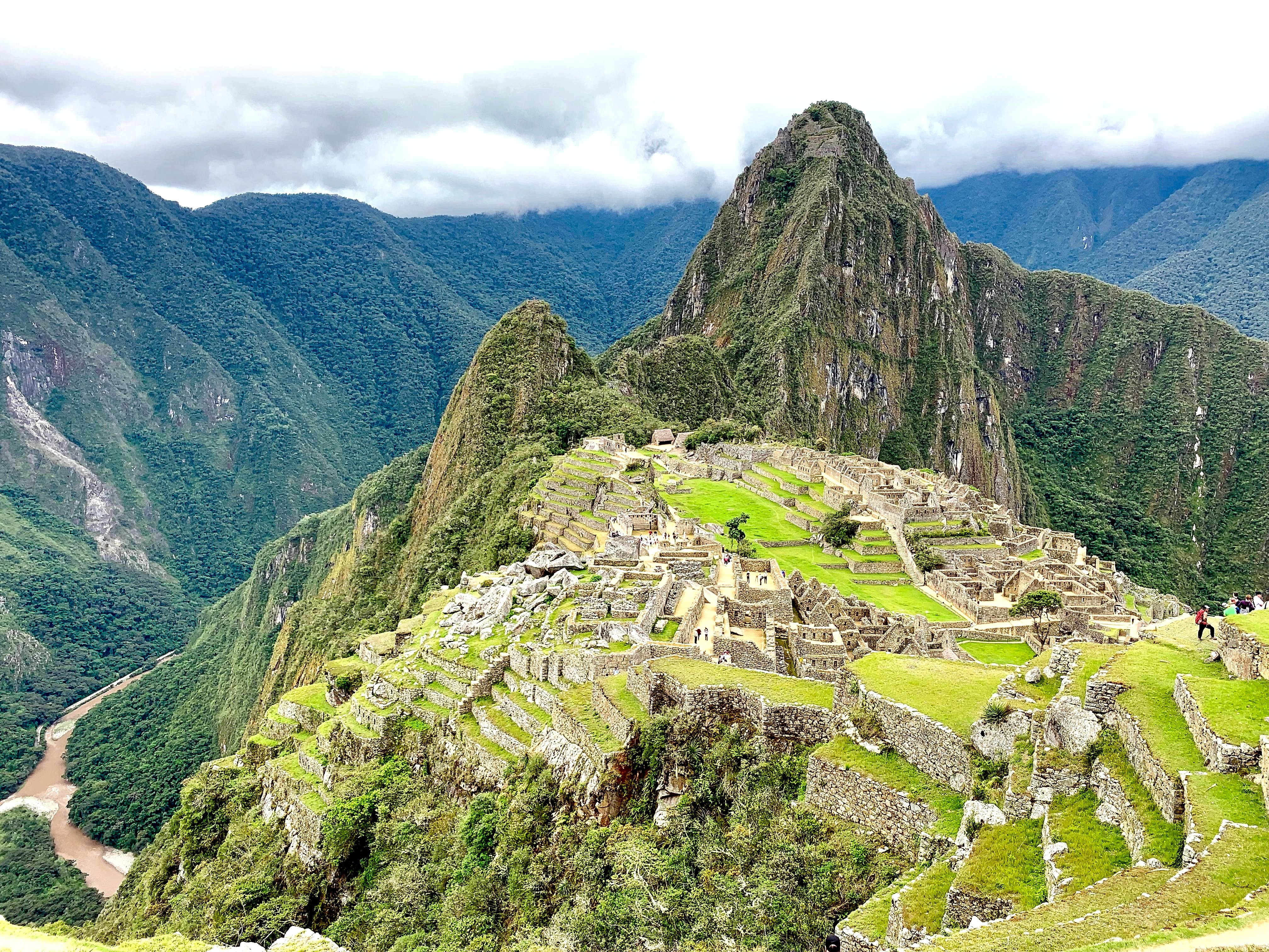 Panoramic view of Machu Picchu ruins and terraces in the Andes, with a steep green mountain and cloud cover behind them.