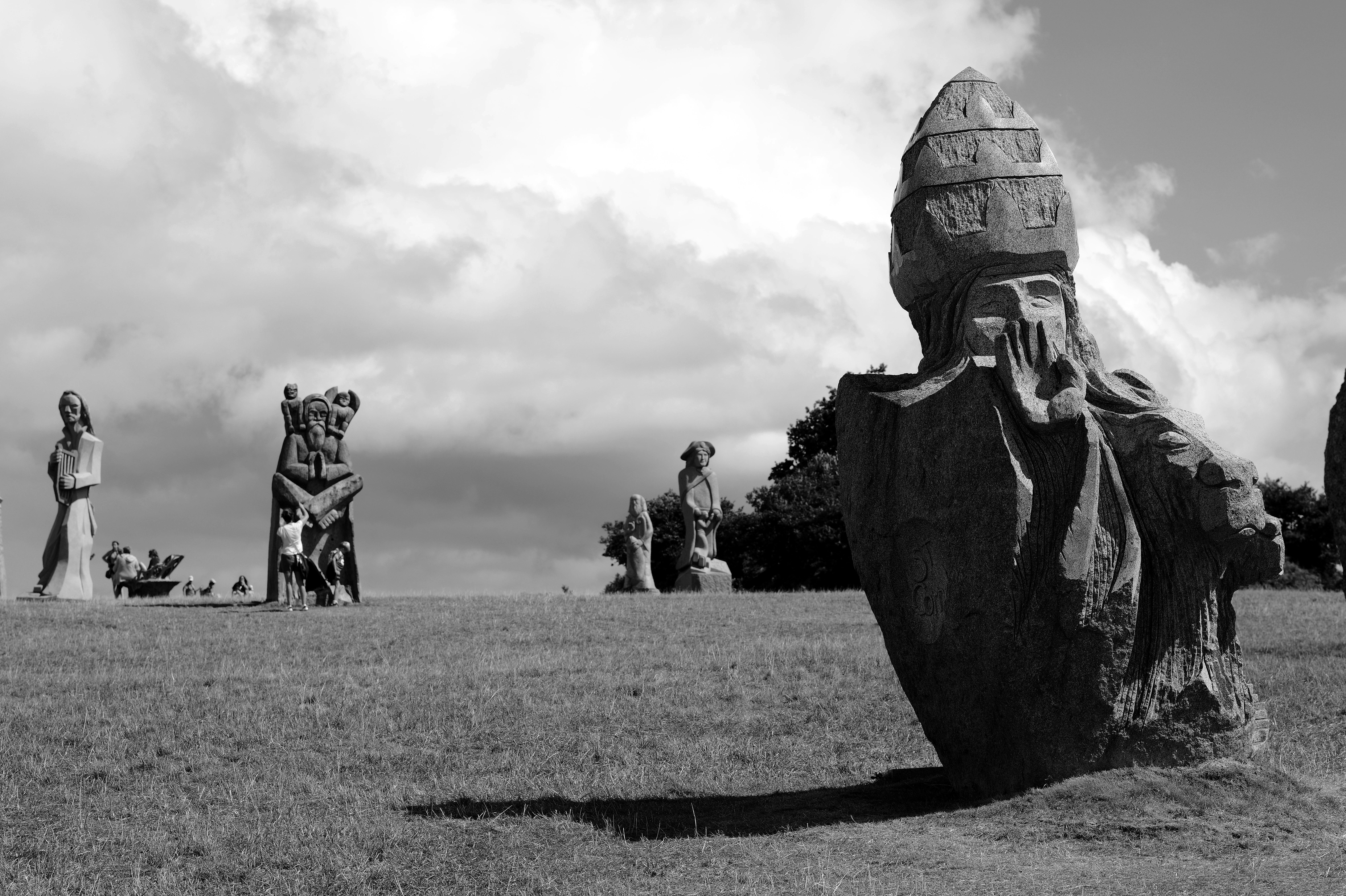 Black-and-white photograph of large stone sculptures in a grassy field with visitors and dramatic clouds at Vallées des Saints in Brittany.