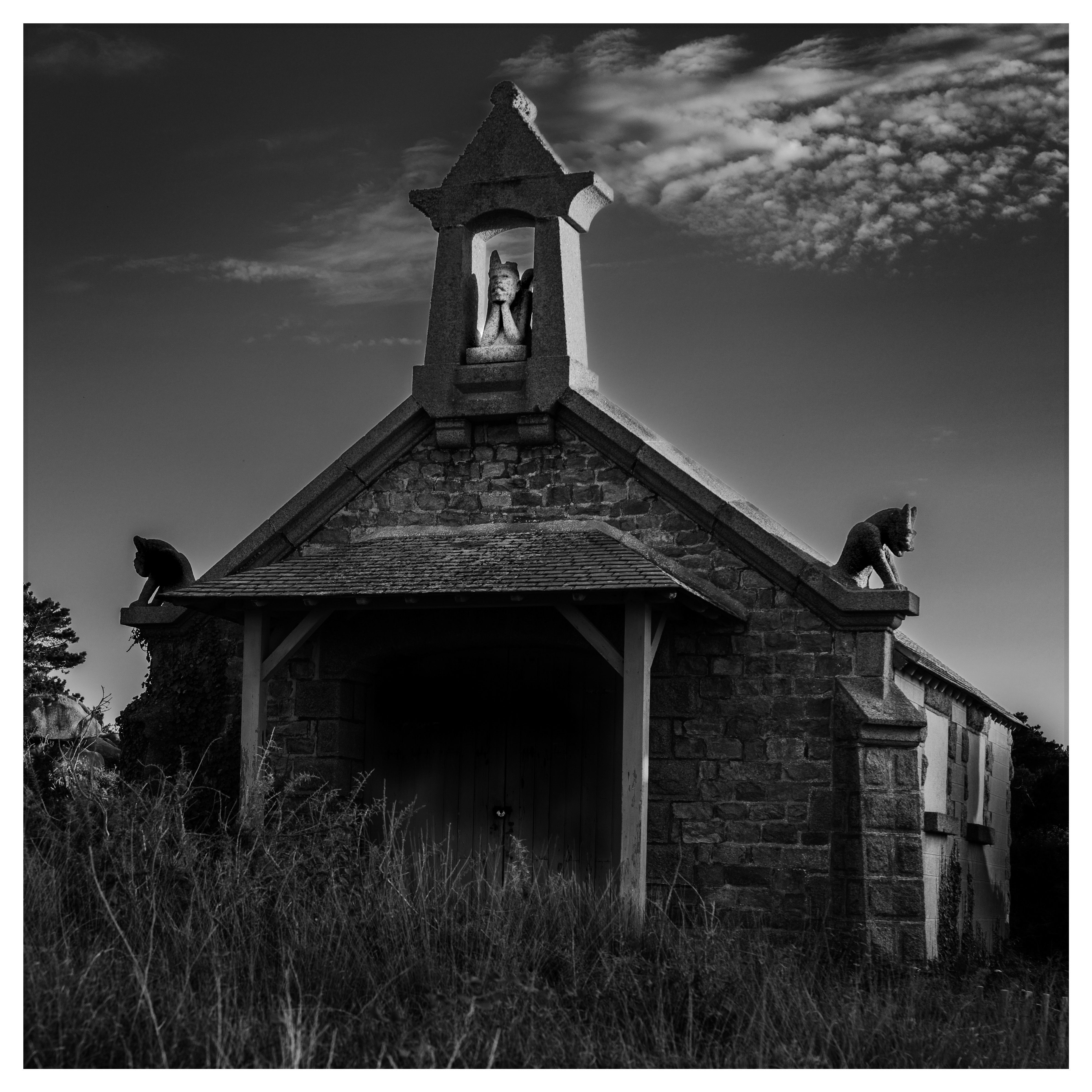 Black and white photograph of a small stone chapel with sculpted roof ornaments and a dramatic sky.