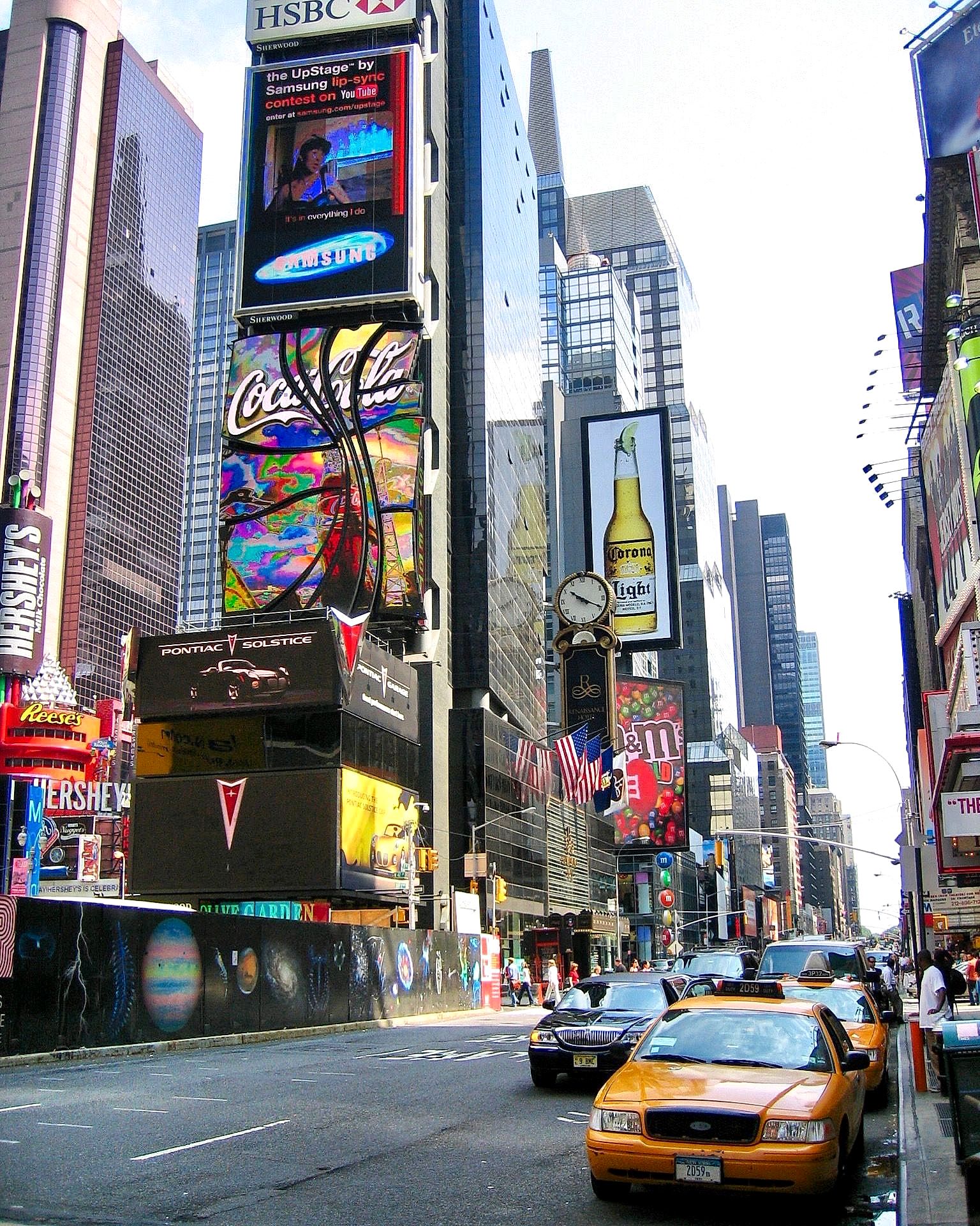 Times Square street scene with stacked advertising screens, skyscrapers, and yellow taxis in bright daylight.