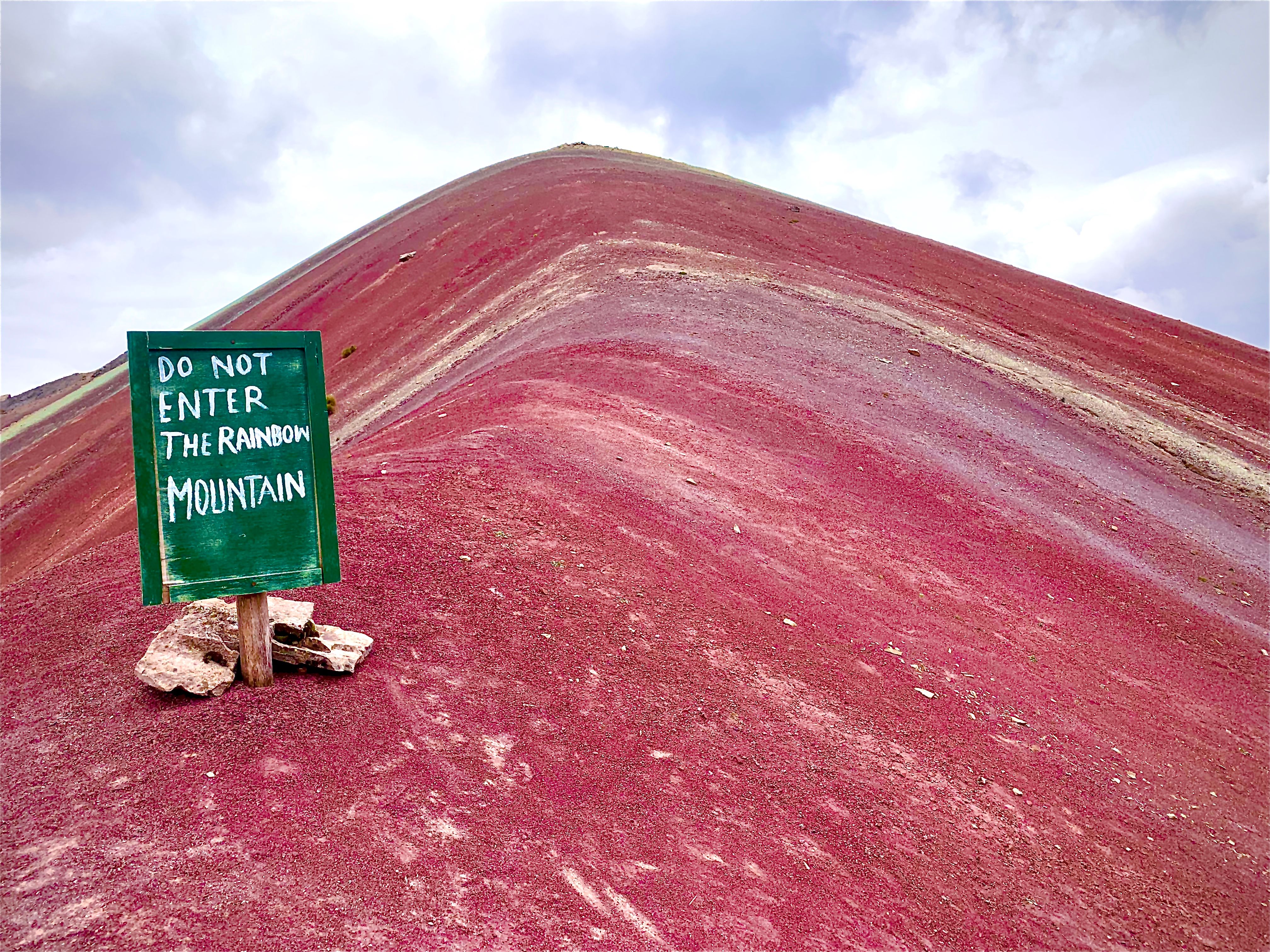 A green sign reading 'Do not enter the Rainbow Mountain' stands beside a steep red-pink mountain slope under cloudy sky.