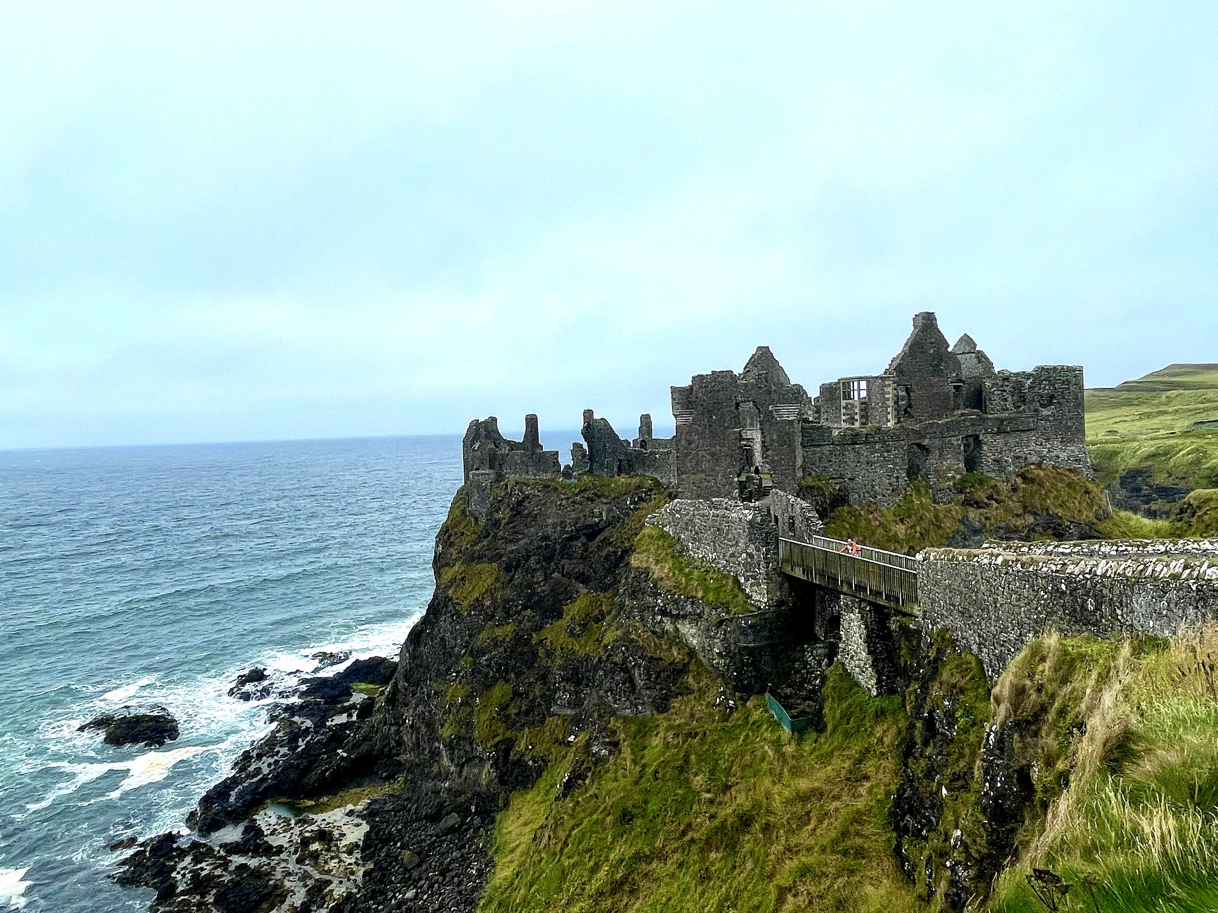 Ruined coastal castle on a cliff above the sea with a footbridge and grassy headland.