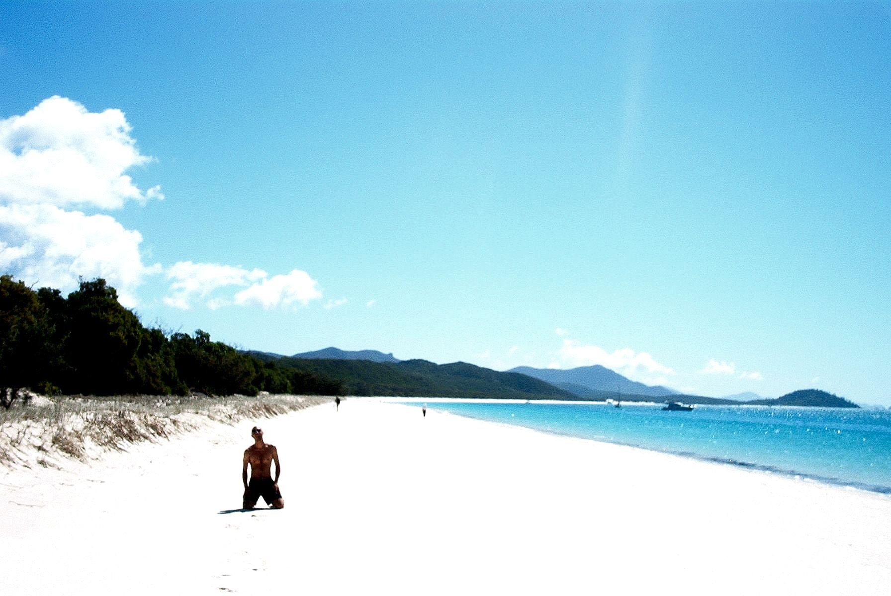 A lone man kneels on a brilliant white sand beach beside turquoise water and green hills under a clear blue sky.
