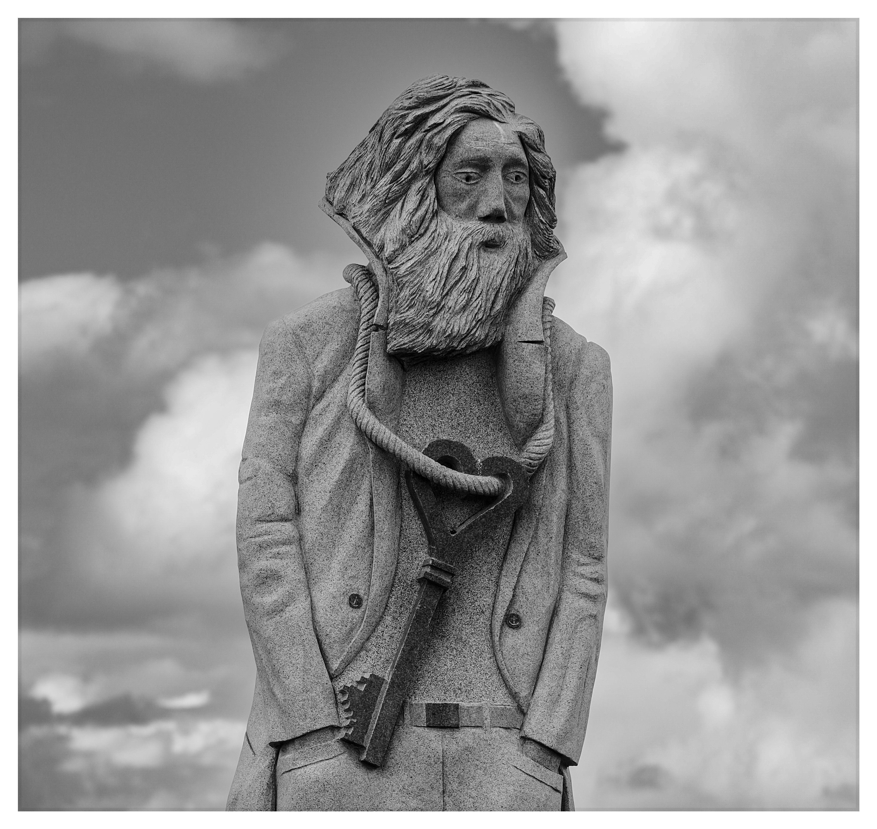 Black-and-white view of a monumental stone saint statue with a beard and long hair, framed against dramatic clouds.