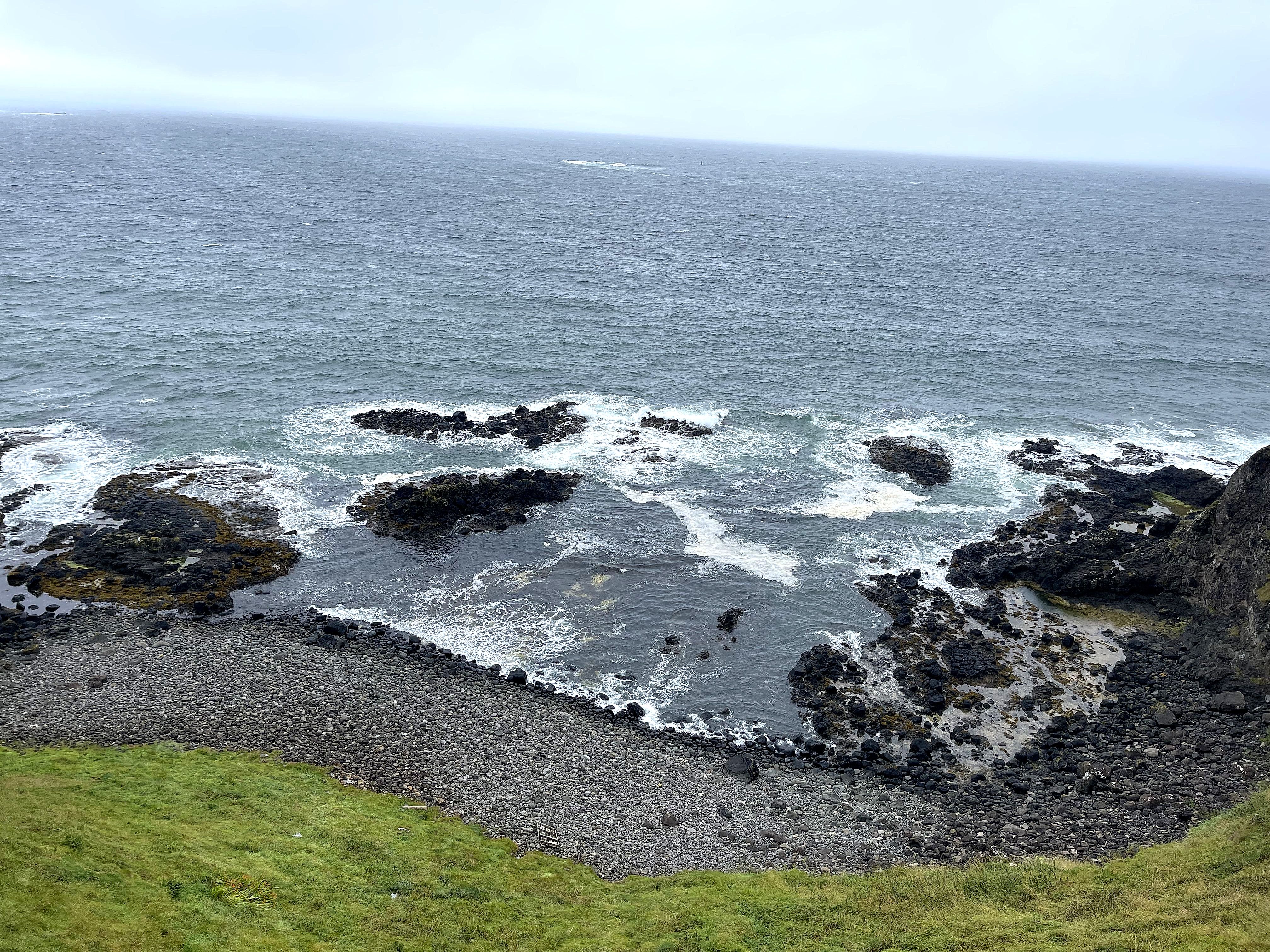 Wide view of a rocky seacoast with waves washing around dark rocks under a misty grey sky.