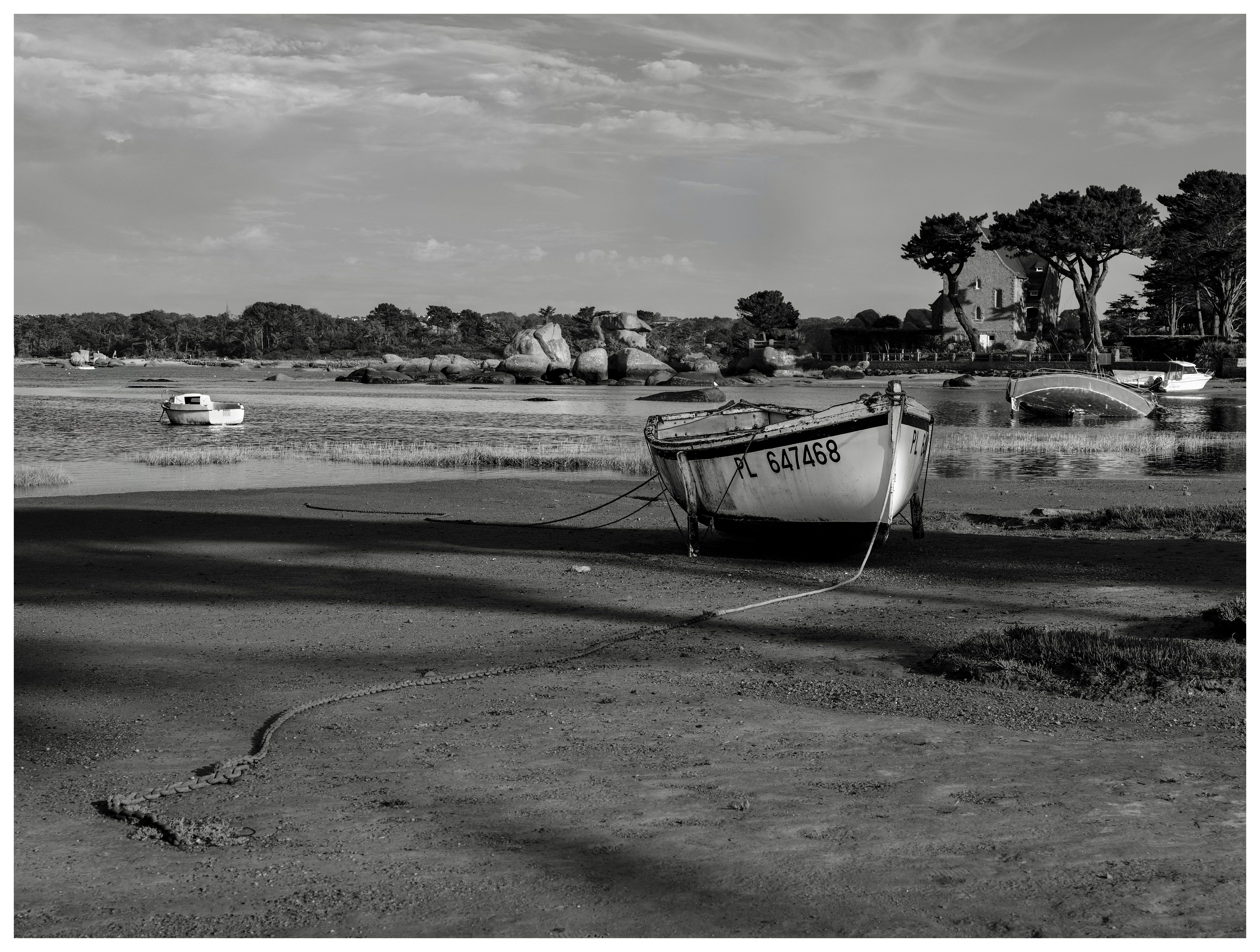 Black-and-white view of small boats resting at low tide on a rocky Breton shore, with a house and trees on the right and open water and clouds beyond.