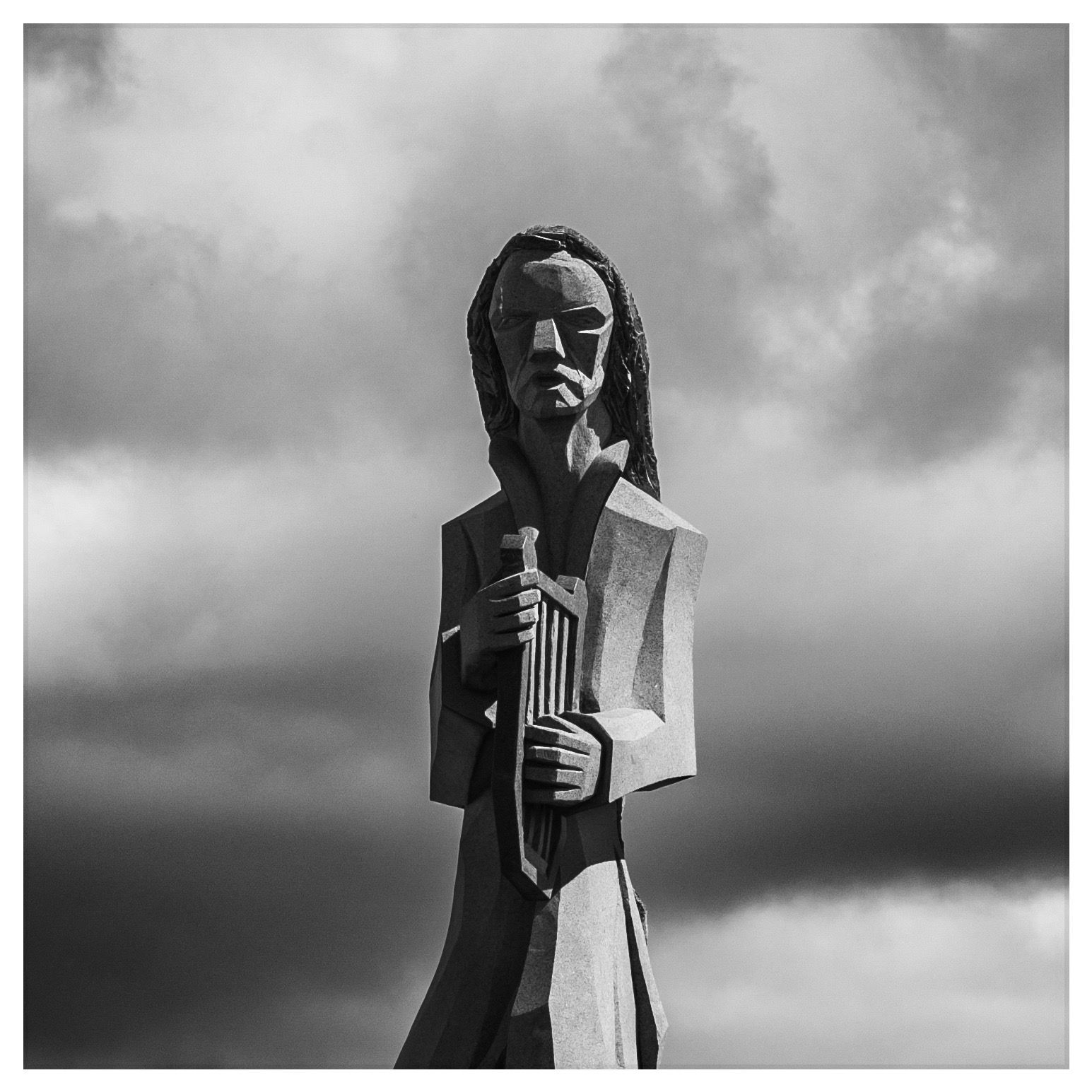 Black-and-white photograph of a tall carved stone statue holding an instrument, set against dramatic clouds.