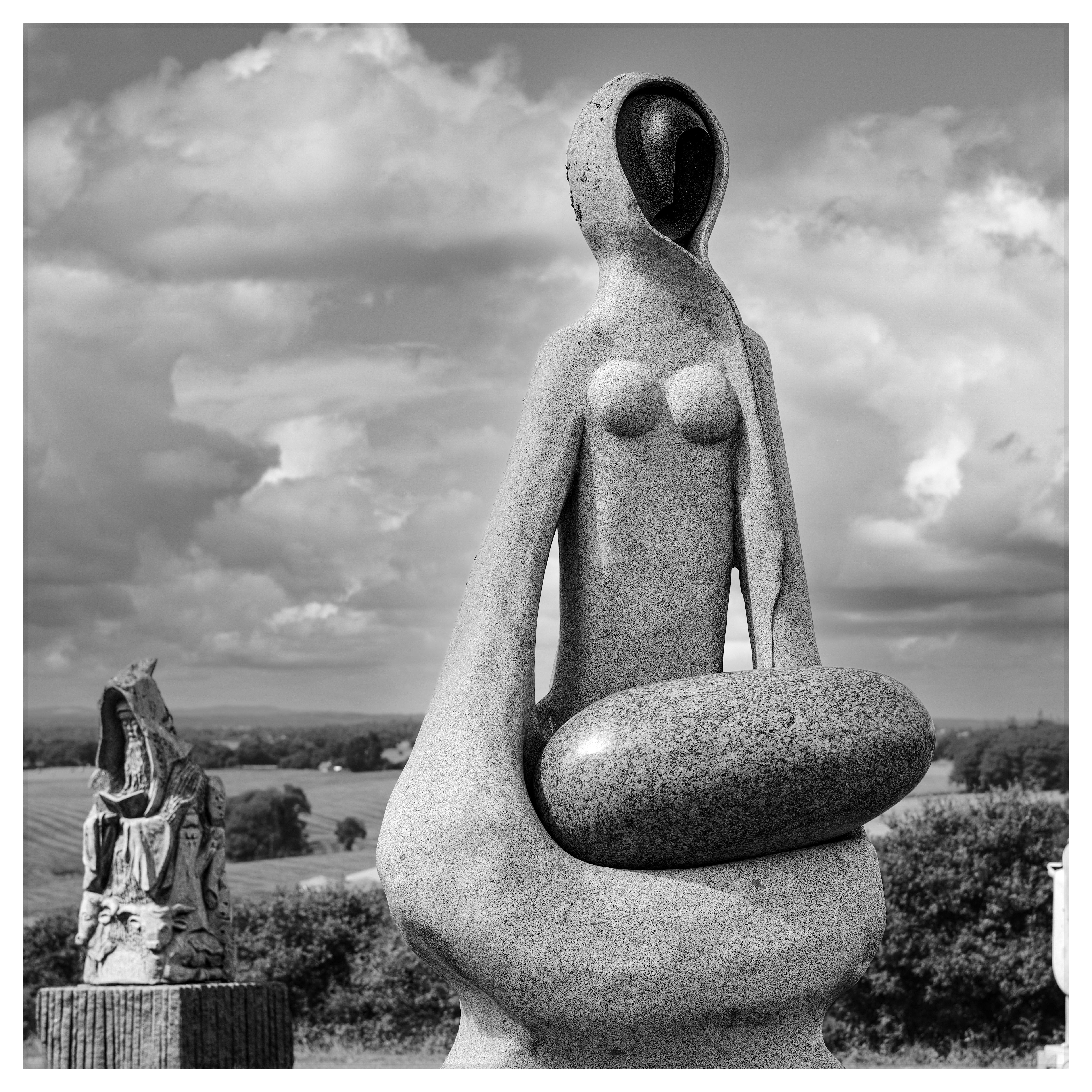 Black and white photograph of a monumental stone sculpture in the Vallée des Saints, with another statue in the background and a cloudy sky.