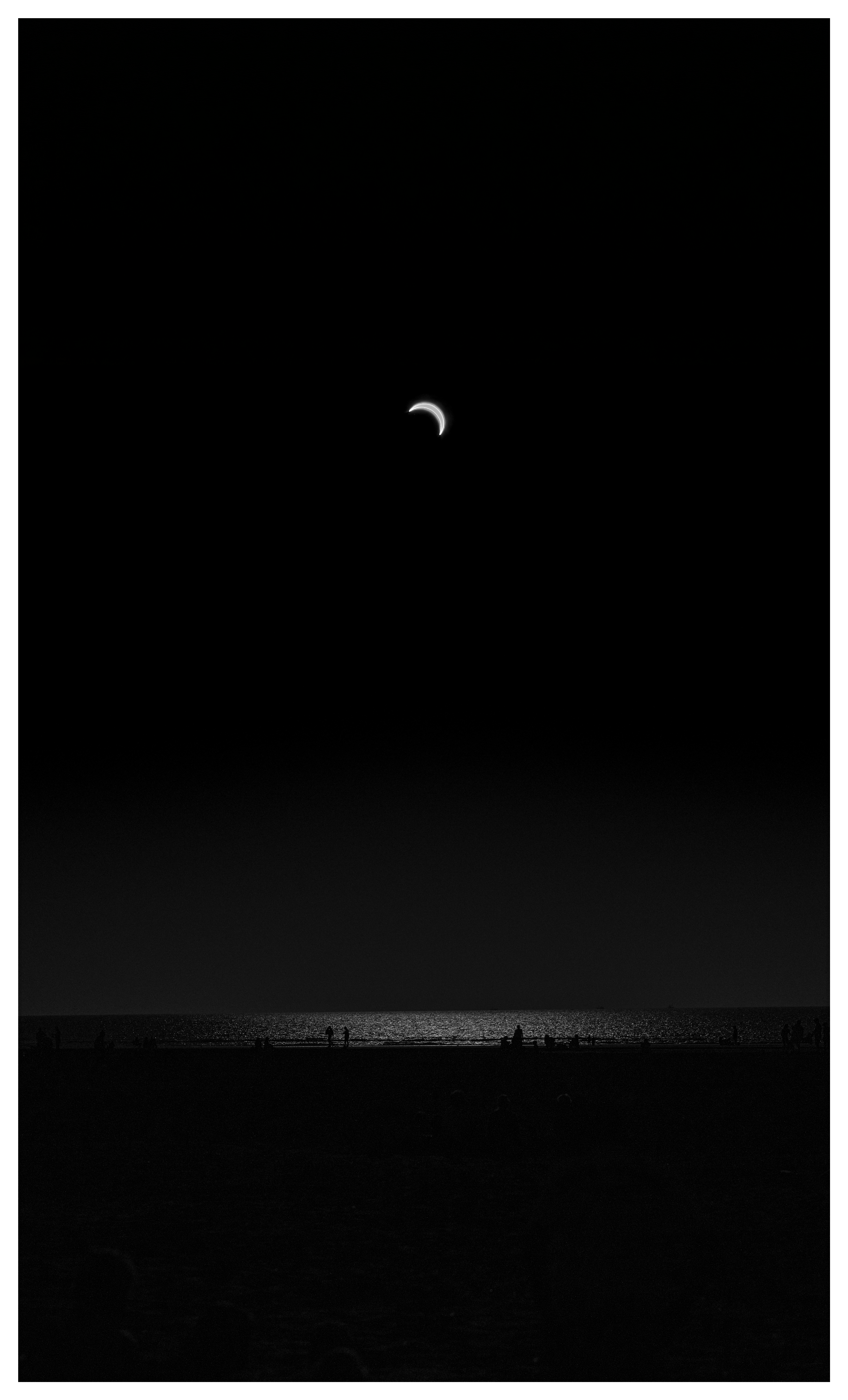 Black-and-white beach scene with a small crescent sun high in a dark sky and tiny figures silhouetted along the shoreline.