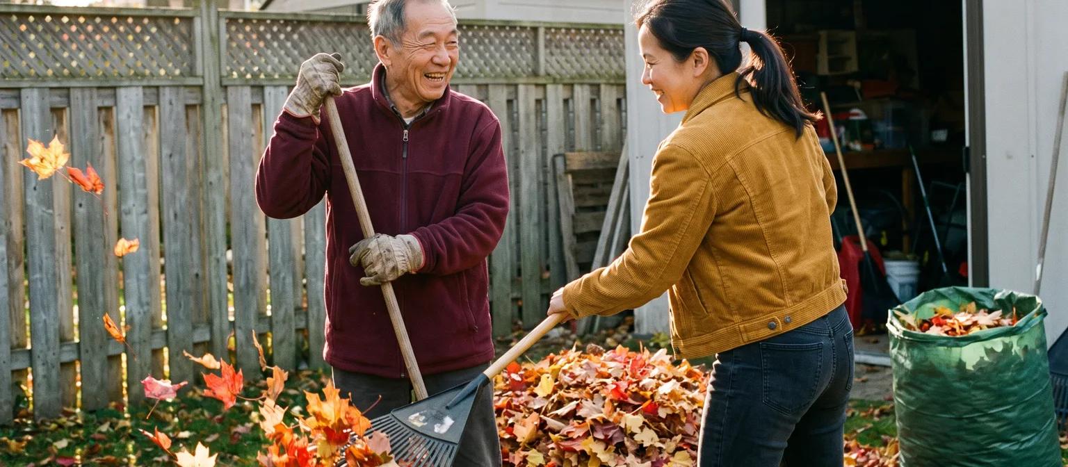 Adult daughter and her father in his late 70s laugh while raking autumn leaves together in the backyard