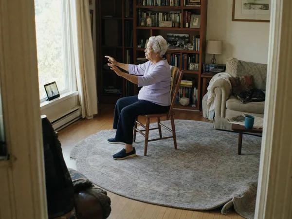 A woman in her late 70s does seated tai chi from a dining chair in her bright living room, following a tablet on the sill