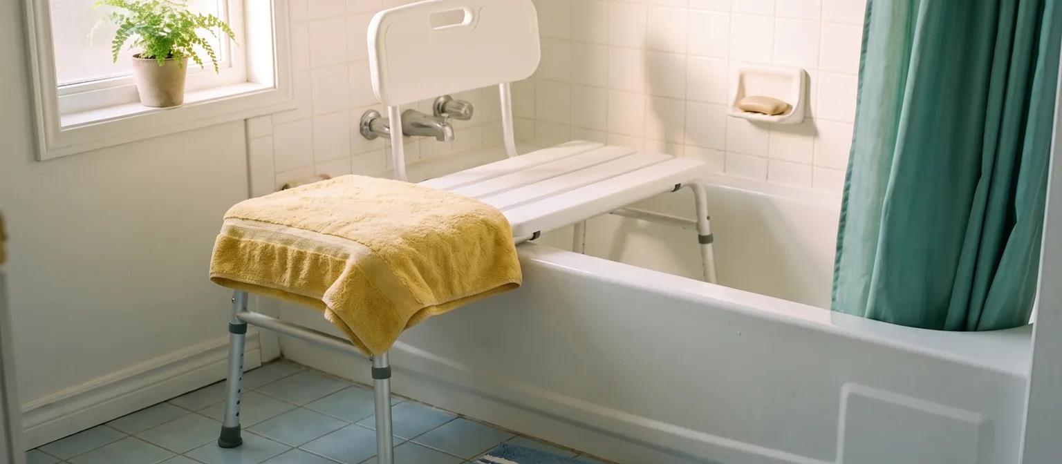 A white tub transfer bench straddles a bathtub rim in a sunny tiled bathroom, two legs in the tub and two on the floor