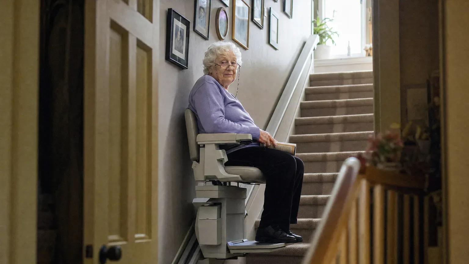 Senior woman seated on a stair lift at the bottom of a carpeted staircase lined with family photos