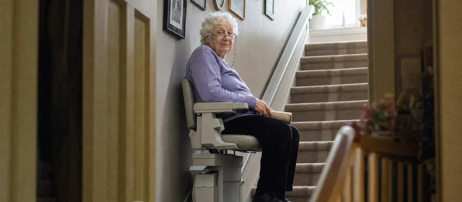 Senior woman seated on a stair lift at the bottom of a carpeted staircase lined with family photos