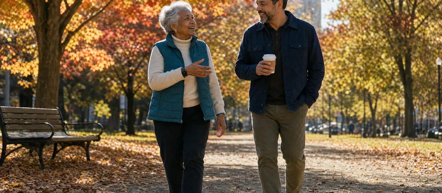 Woman in her late 70s walks a sunny autumn park path with her son, a small black alert device on her wrist