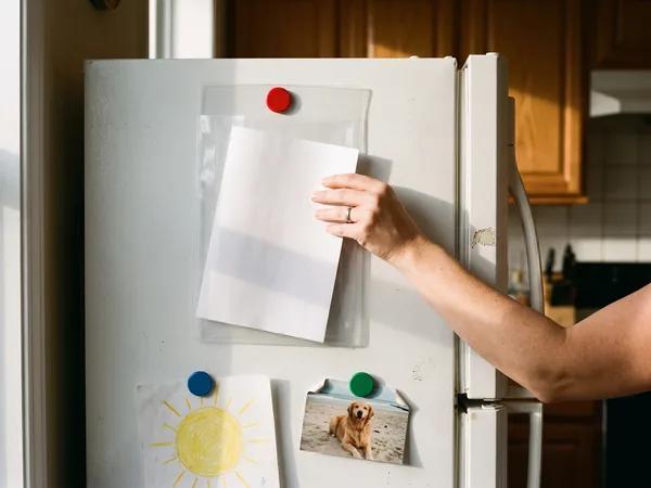 A hand slides a folded document into a clear sleeve on the fridge door, where it is easy to find in an emergency