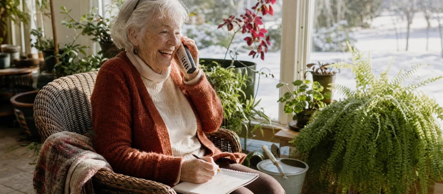 A woman in her late 70s laughs on a call in her sunny plant-filled sunroom, pencil and notebook on her knee.