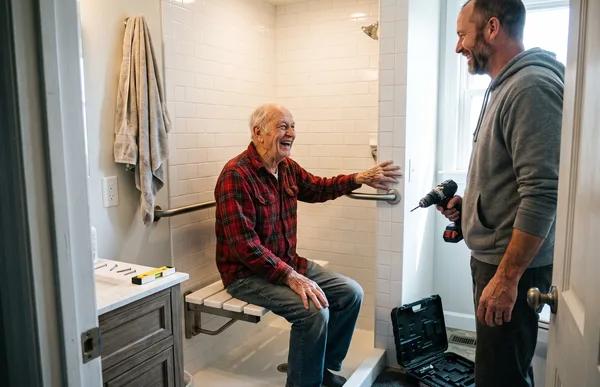 A man in his 80s laughs from a new fold-down shower bench, hand by the grab bar, as his son holds the drill