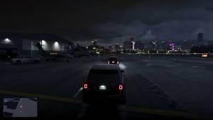 Gameplay of an SUV following another across an airfield at night, with private jets by a hangar and the Vice City skyline behind.