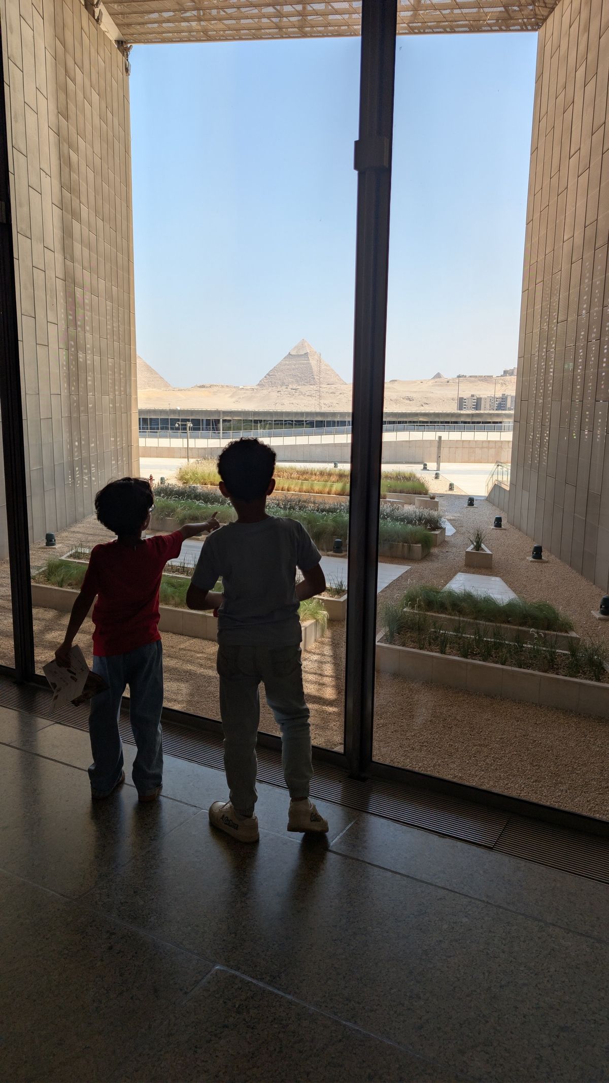 Two children looking through a window towards the Pyramids of Giza.