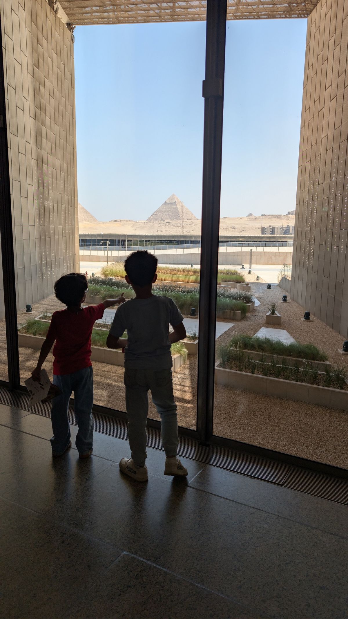 Two children looking through a window towards the Pyramids of Giza.