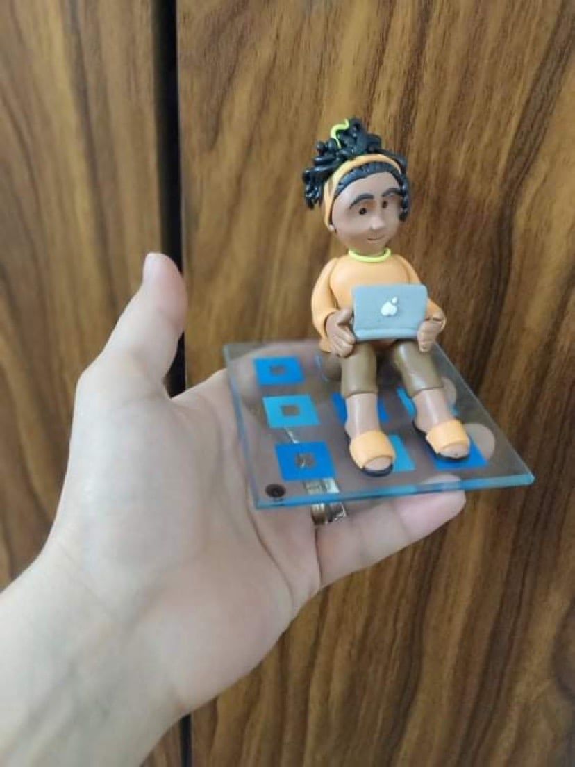 The finished clay figure holding a tiny laptop, resting on its glass base in the palm of a hand.