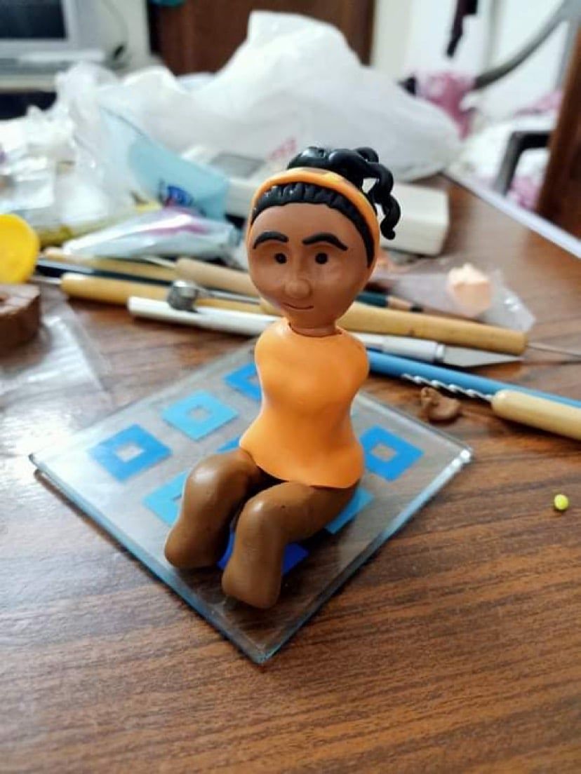 A handmade clay figure in an orange top sitting on a small glass base, surrounded by sculpting tools on a wooden table.