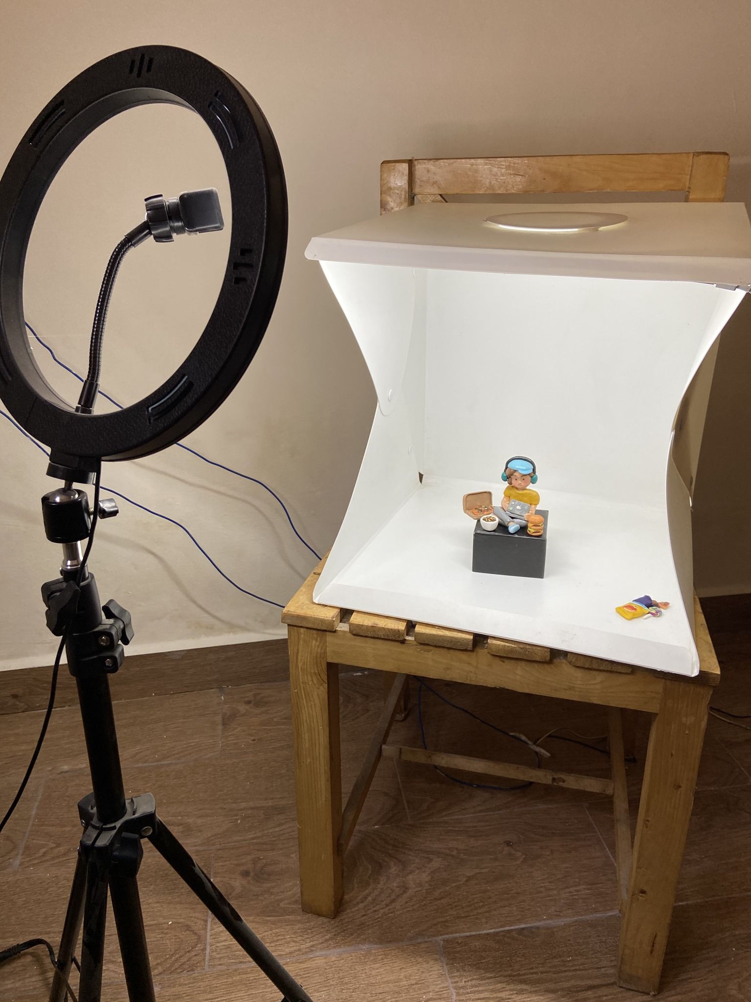 A ring light on a tripod beside a small white lightbox on a wooden chair, with a clay figure posed on a black stand inside.