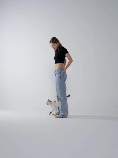 a woman in a black crop top and blue jeans is standing next to a cat .