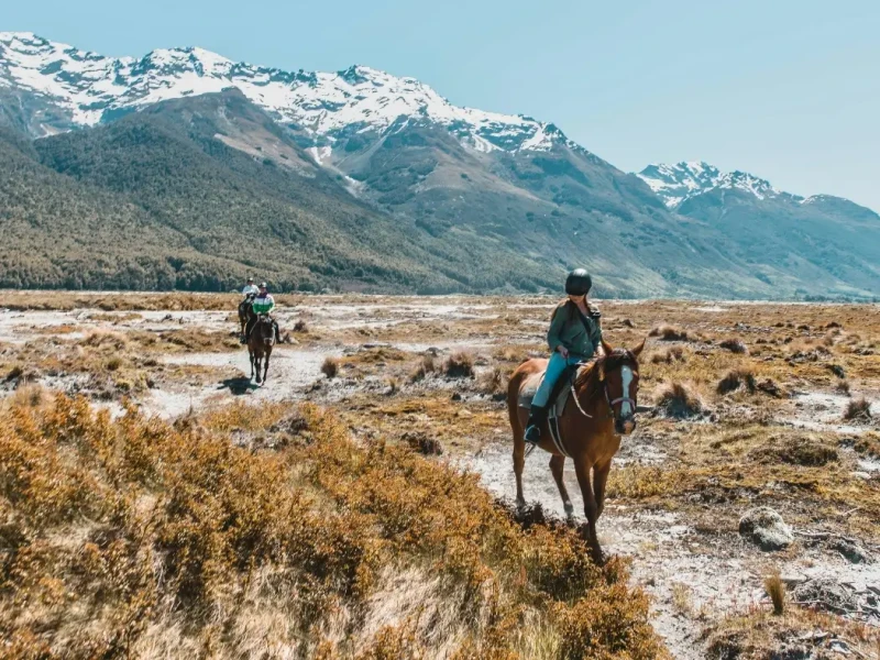 Travellers horse riding through an open South Island valley with snow-capped mountains in the distance, highlighting outdoor adventure, scenic landscapes and non-ski winter activities to add to a New Zealand travel itinerary.