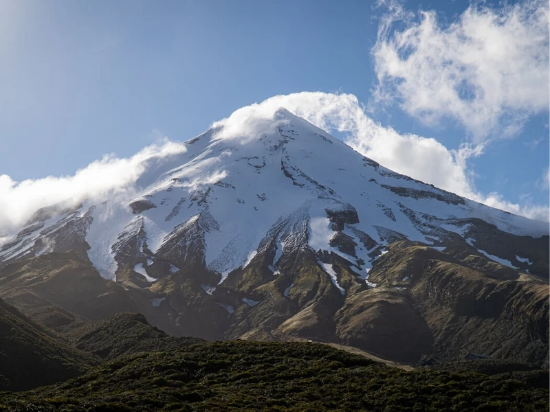 A close view of snow-covered Mount Taranaki with clouds around the summit and native bush below, highlighting one of New Zealand’s most iconic volcanic regions for North Island hikes and outdoor adventure travel.