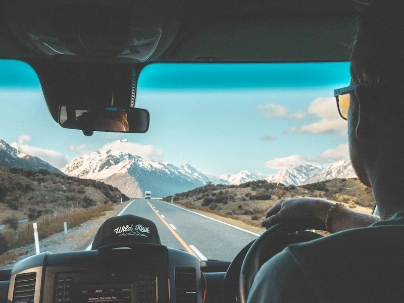 A photo through the windshield of a Wild Kiwi premium New Zealand tour van looking out at snow-capped mountains and a straight highway.