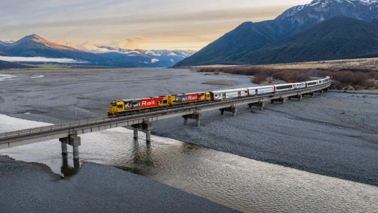 A scenic view of New Zealand’s TranzAlpine train crossing a long bridge over a braided river in the South Island, surrounded by wide alpine valleys, rugged mountains and soft morning light. This dramatic TranzAlpine rail journey scene captures the beauty of one of the world’s most scenic train rides, travelling through the South Island between Christchurch and Greymouth.