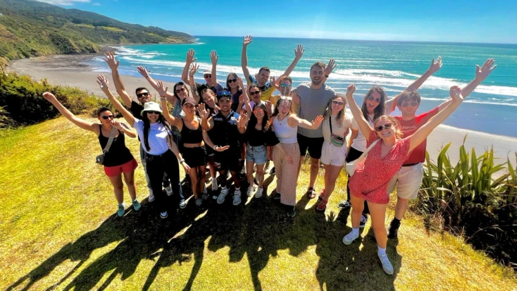Group of travellers smiling and raising their hands on a cliff above a turquoise New Zealand beach during a Wild Kiwi road trip adventure.