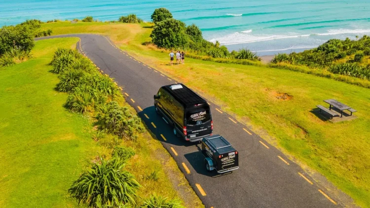 Wild Kiwi van driving along a coastal New Zealand road near Raglan with turquoise ocean waves and travellers enjoying the view — promoting the Wild Kiwi Northern Voyager trip giveaway.
