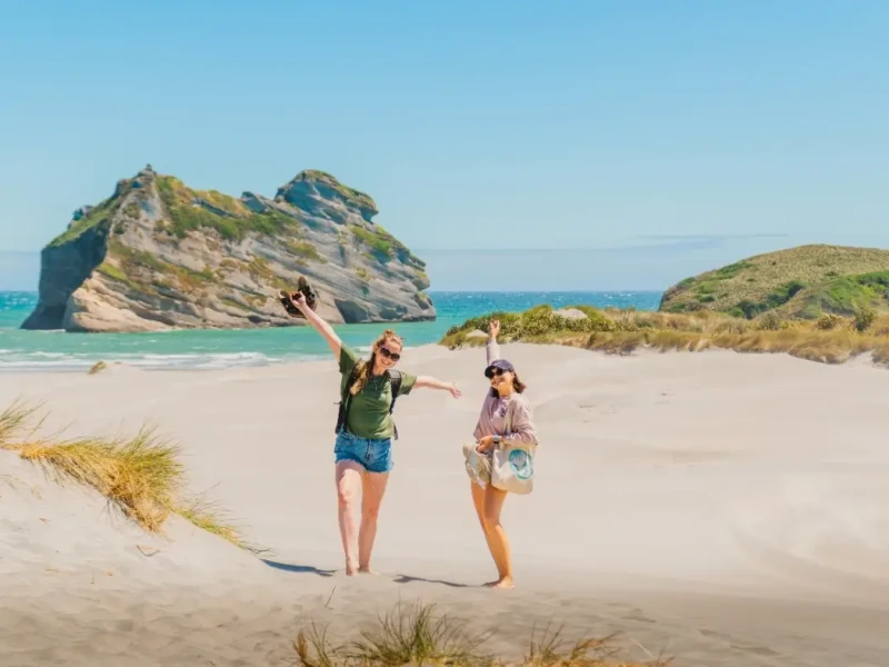 Two female solo travellers pose happily on sand dunes with a backdrop of the sea at New Zealand's famous golden bay while on a small group adventure tour in Spring.