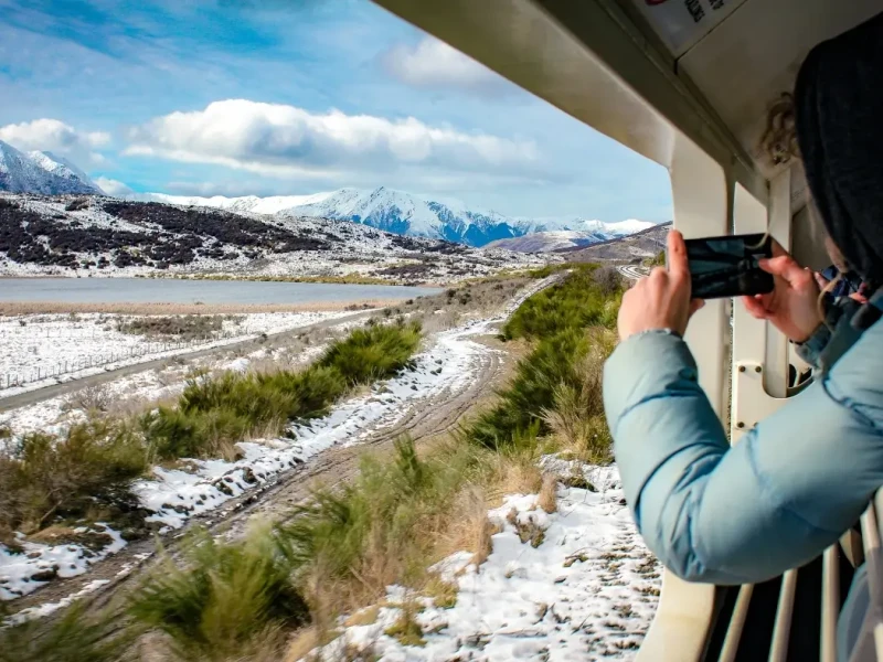 A passenger photographs snowy South Island scenery from the open-air viewing carriage of the TranzAlpine train in New Zealand. Surrounded by mountain ranges, winter grassland and a lakeside landscape, this image shows the immersive outdoor viewing experience that makes the TranzAlpine one of New Zealand’s most scenic train journeys.